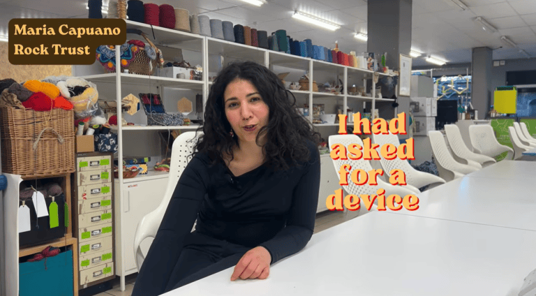 A person sits at a table as though talking to the camrera. they are in an office with sewing equipment.